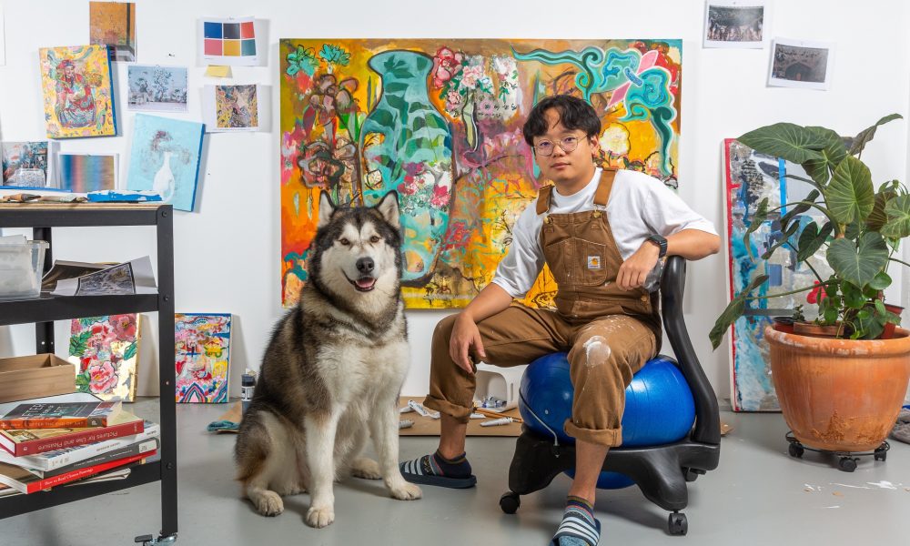 Image: Xuanlin Ye with his companion, a husky named Baobao, MANA Contemporary, Chicago, 2022. He is seated on a blue exercise ball. Behind him is one large colorful painting and surrounding that are many smaller pieces of paintings and reference images on the wall. To his right is a plant in a large terracotta pot. Photographed by Guanyu Xu. Image Courtesy of the artists.  