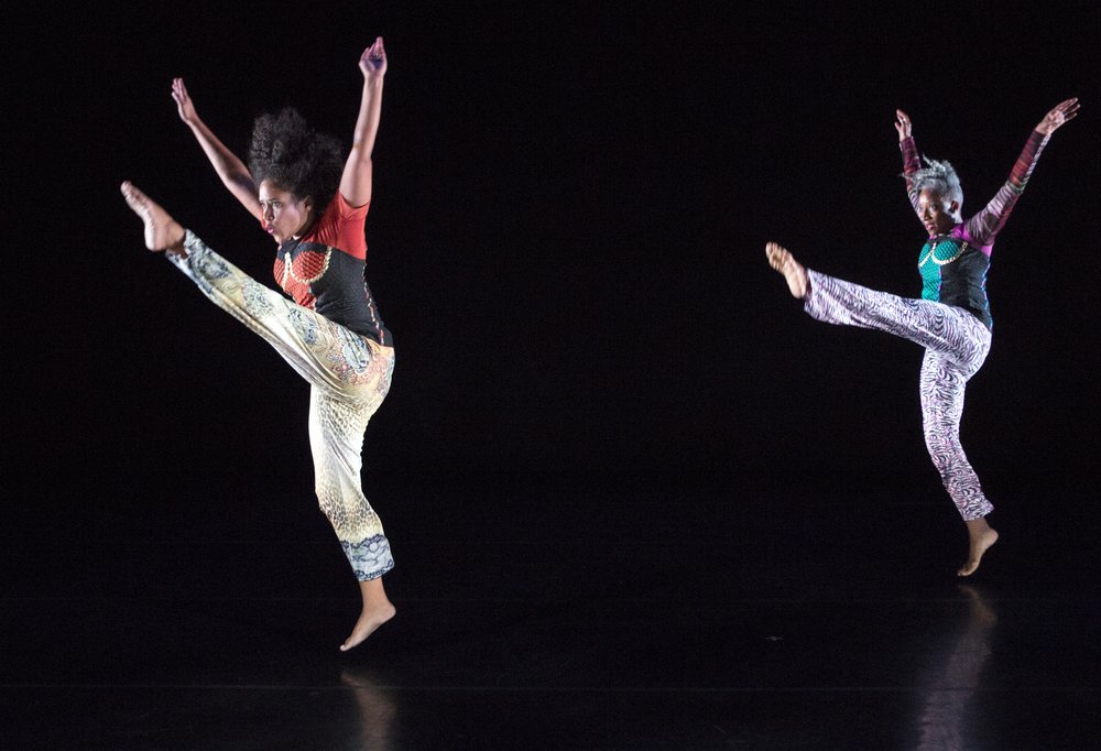 Image: From the production BOOM!. Two dancers captured mid-dance, facing the left of the image with their arms raised and their left legs in the air, on a dark, black stage and a dark, black background. On the left, Leslie Cuyjet wears light colored pants and a red and black top. On the left, Cynthia Oliver wears light purple pants and a black and teal top. Photo by Photo by Yi-Chun Wu, courtesy of Cynthia Oliver.