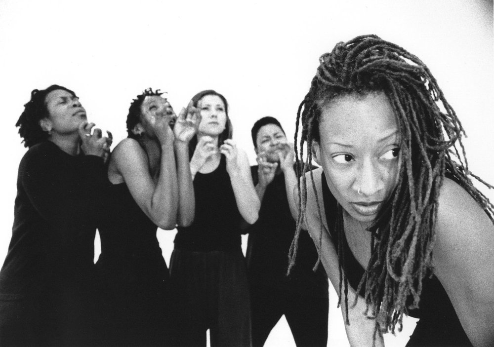 Image: From the production SHEMAD. A black and white photo of Cynthia Oliver (foreground, right) with four dancers behind her. All dancers are wearing black pants and black tops with different iterations of sleeves; their hands are lifted toward their faces and their eyes are closed. Oliver is bending forward, wearing a black tank top and looks out of the picture plane to the right. She has long, thin dreadlocks that fall over her shoulder. Photo by Andrew Eccles, courtesy of Cynthia Oliver.