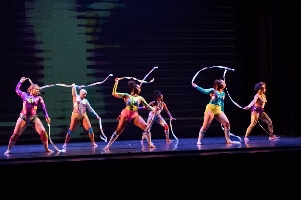 Image: From the production Tether. Six women are dancing on stage with some sort of ribbon or tethering material in their hands. Some wear knee pads and others wear knee-high socks. They are all wearing very brightly colored and shiny costumes of shorts and long sleeve shirts. Behind them is an abstract animation/visual. Photo by Natalie Fiol, courtesy of Cynthia Oliver.
