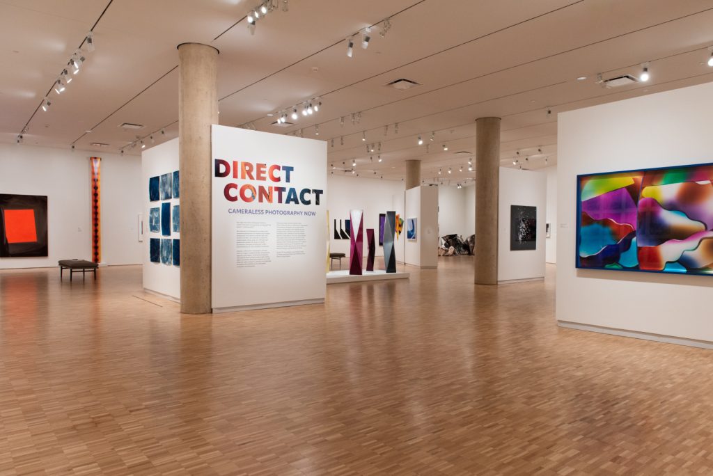 Image: Installation view, Direct Contact: Cameraless Photography Now, 2023. A floating wall with text reading Direct Contact stands in the center, in a gallery space with colorful artworks. Courtesy of Sidney and Lois Eskenazi Museum of Art, Indiana University. Photo: Shanti Knight.