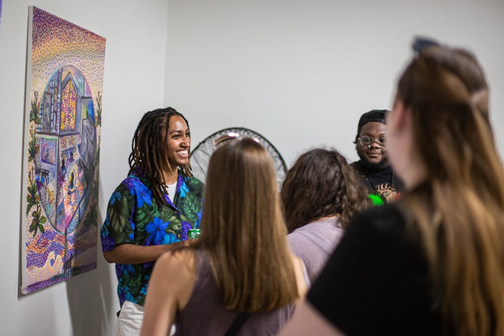 Image: Maria Lavender is smiling toward a crowd of people while standing in front of a collaborative painting. The painting depicts a distinct and impressionistic world made up of various shades of blues, violets, and yellows within large circles surrounded by natural environments. The work on display is part of a collaborative exhibition put on by PGOBA as part of CANJE. Photo by Alexa Cary.