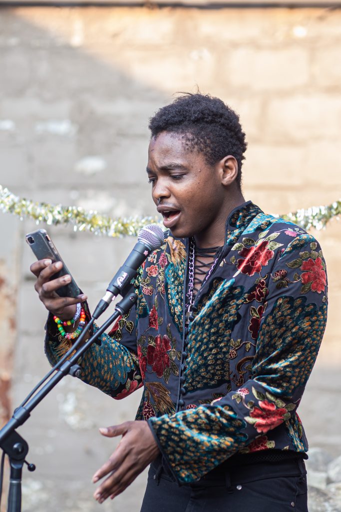 Image: Ezra Collom sings while wearing a dark velvet shirt. Their hand is gesturing to the left. Photo by Alexa Cary.