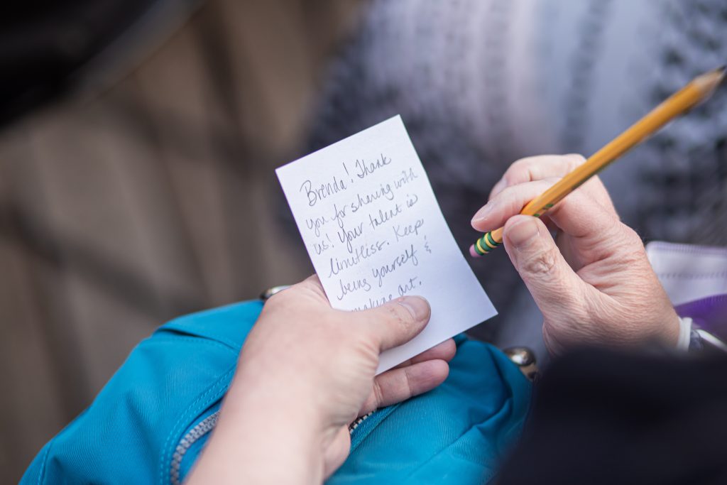 Image: A close up of two hands: one holding a pencil and the other holding a piece of paper for writing their feedback to the performances. The piece of paper says, "Brenda! Thank you for sharing with us! Your talent is limitless. Keep being yourself & making art." Photo by Alexa Cary.