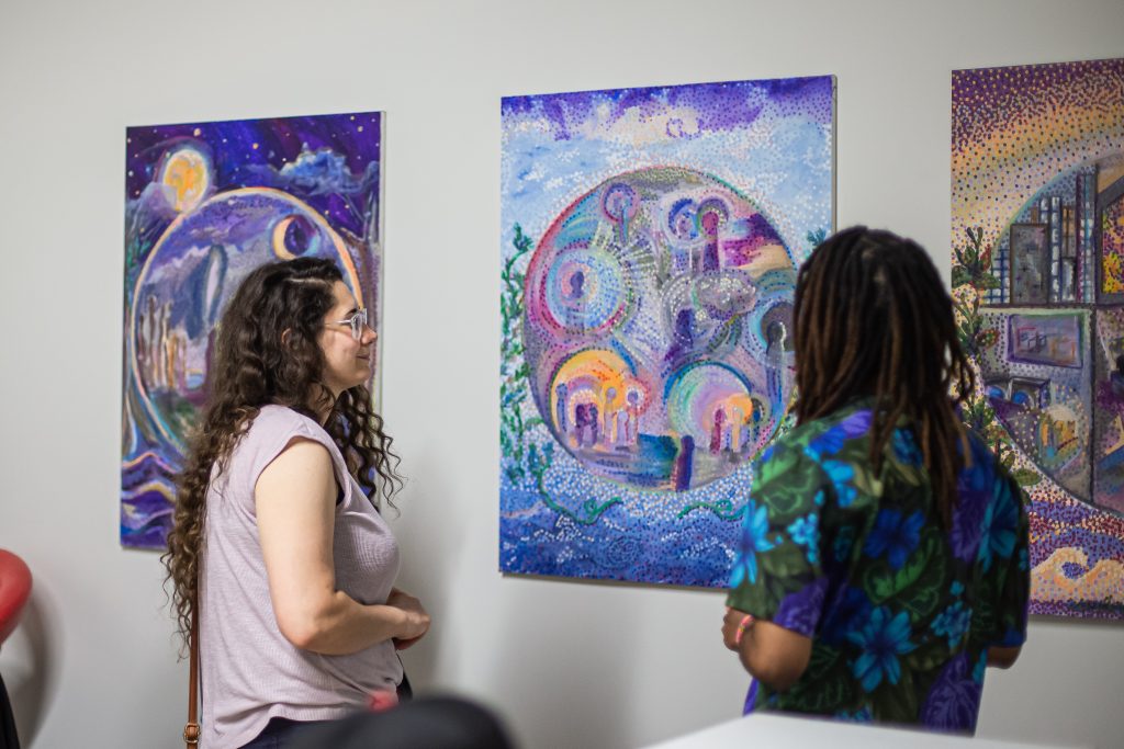 Image: Maria Lavender stands with an audience member in front of a series of three colorful collaborative paintings.  Each painting depicts distinct and impressionistic worlds made up of various shades of blues, violets, and yellows within large circles surrounded by natural environments. Photo by Alexa Cary.