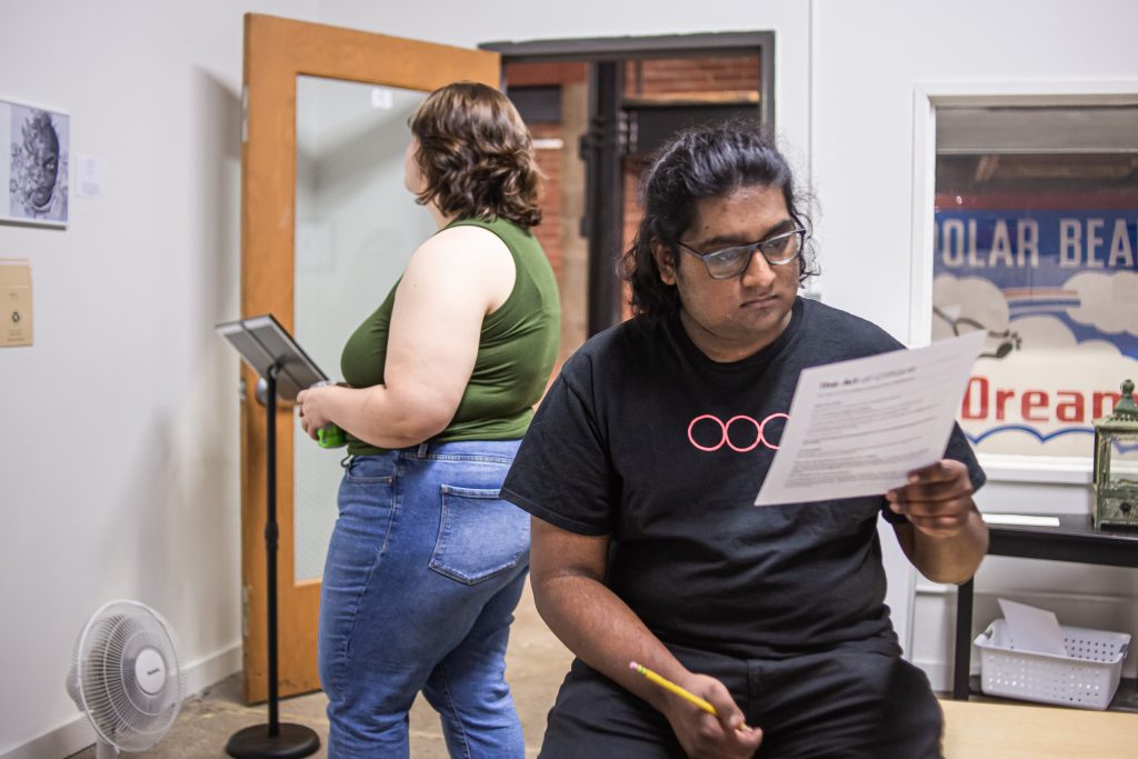 Image: Two people stand in a gallery viewing the collaborative exhibition The Art of Critique, organized by PGOBA as part of CANJE. The person on the right is holding a piece of paper with instructions on how to provide feedback to the artists on display. Photo by Alexa Cary.