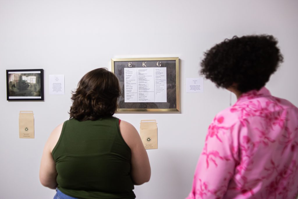 Image: Two people stand in a gallery viewing the collaborative exhibition The Art of Critique on the wall. We can see two artworks on display with little pockets underneath them for folks to place their written feedback. Photo by Alexa Cary.