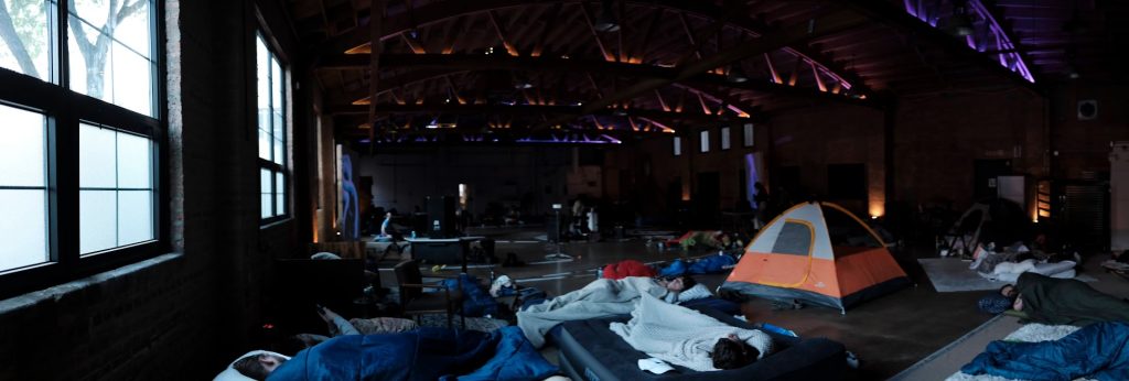 Image: A wide-angle photograph of StretchMetal's Drone Sleepover event. Participants are sleeping on the floor of a long bowtrussed room with bright windows on the left wall. The participants are wrapped in blankets, and there's one orange tent on the right. Image courtesy of StretchMetal. 