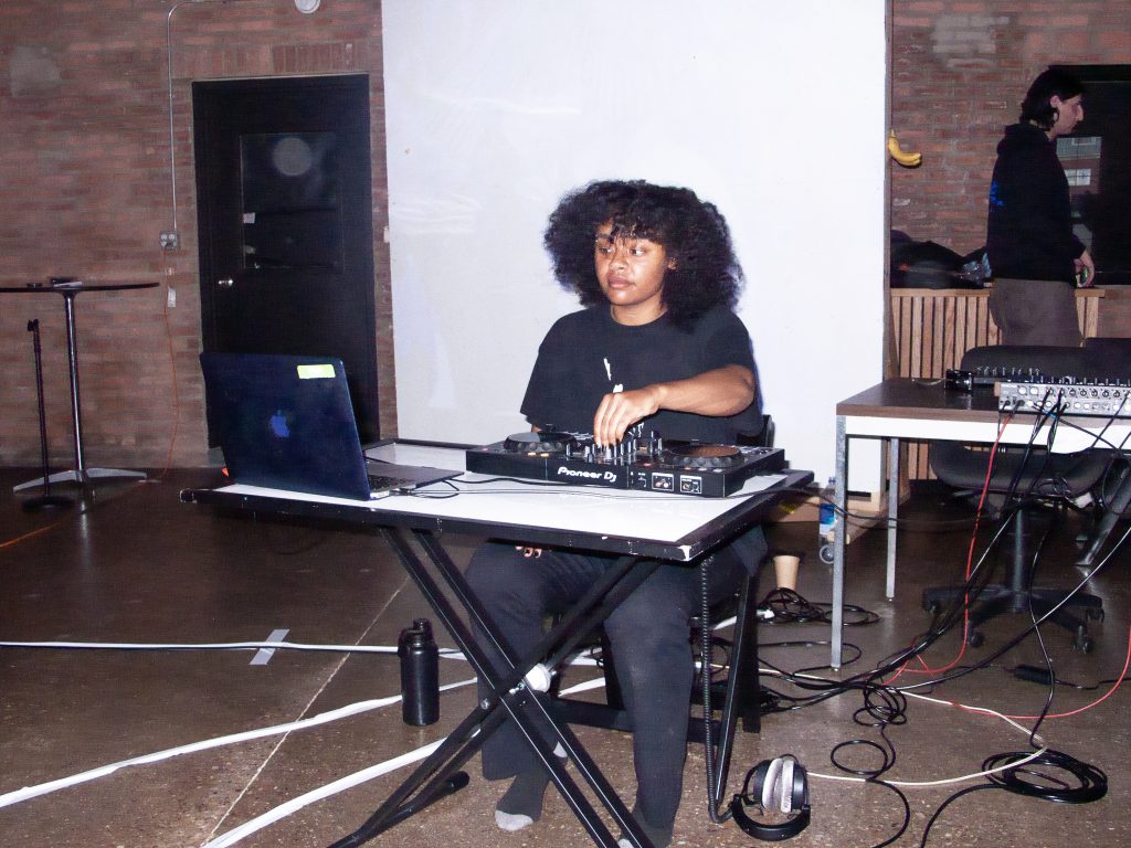 Image: The musician Leah Damte seated at a folding desk with a laptop and a small DJ set in front of them. Behind them is a white scrim and to their right is a table covered with sound equipment, running down to a mess of wires beneath them. They play their rooster set at the Drone Sleepover. Photo courtesy of StretchMetal. 