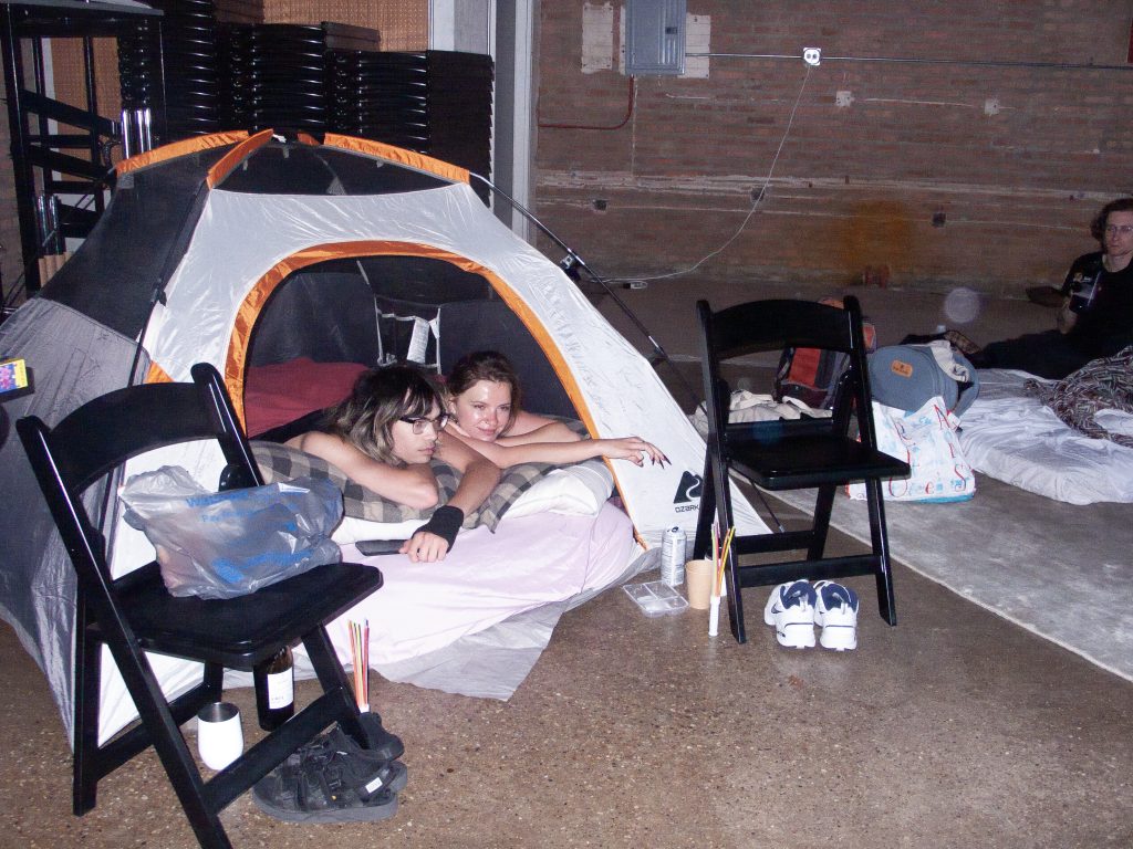 Image: Two participants lay with their faces and arms outside of an open tent. On either side of the tent are two black folding chairs with shoes underneath them. To their right is a carpet with some backpacks and a mattress on it. A person sits on this mattress. Behind all of them is a brick wall. Image courtesy of StretchMetal. 