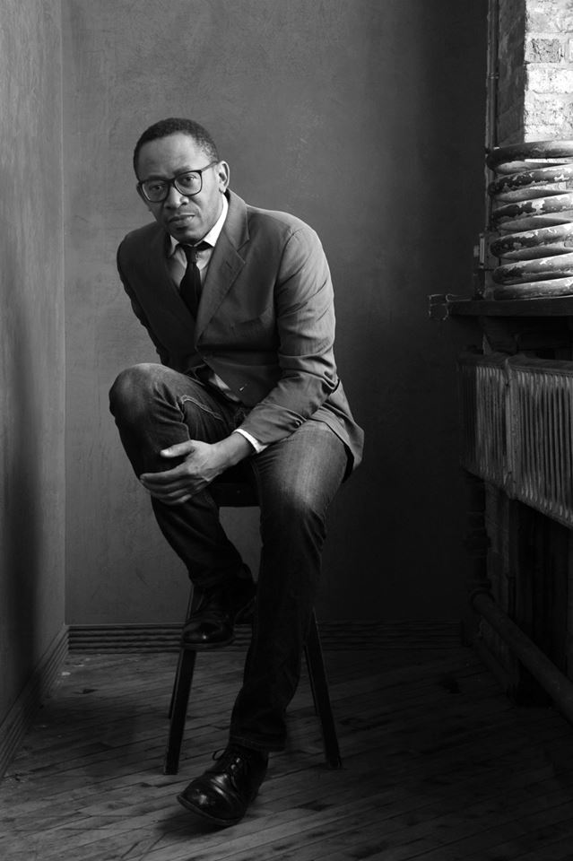 Image: A black and white photograph of Norman Teague. He sits on a chair with one leg resting on the chair and a hand on his leg. Photo by ReportArch/Andrea Ferro Photography. From the article: https://www.nytimes.com/2023/05/20/arts/design/plastic-venice-biennale.html