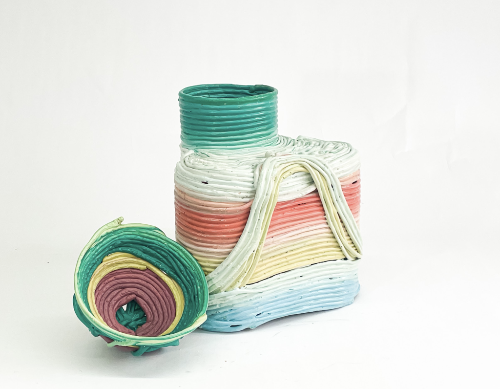 Image: A vessel made out of recycled plastic by Norman Teague. The plastic is worm-like and molded together in strips. It gradiates in color from a light blue to off-white, to yellow to salmon, back to white then to green. The vessel's cap is off and set to the side. Photo by Norman Teague Design Studios, 2023.