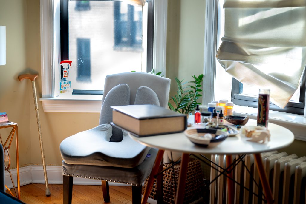 Image: An interior shot of Perry's home. There is a table at the forefront with an assortment of objects including a book, oils, a candle, medicine, and a basket. Next to the table is a chair with extra cushions and a plant. Perry's cane rests in the corner next to a window. Photography by Tonal Simmons. 