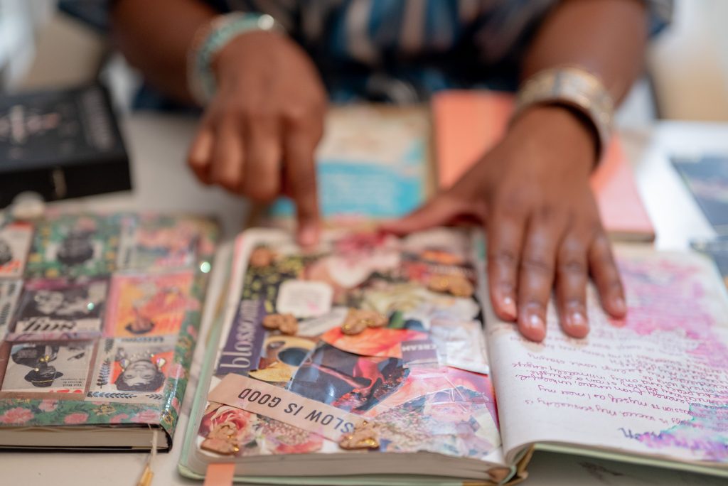 Image: Close up of collaged pages in Perry's journal. Perry's hands are placed on the pages pointing to the lower half of the page. One saying on the page says, "Slow is Good." Photo by Tonal Simmons. 