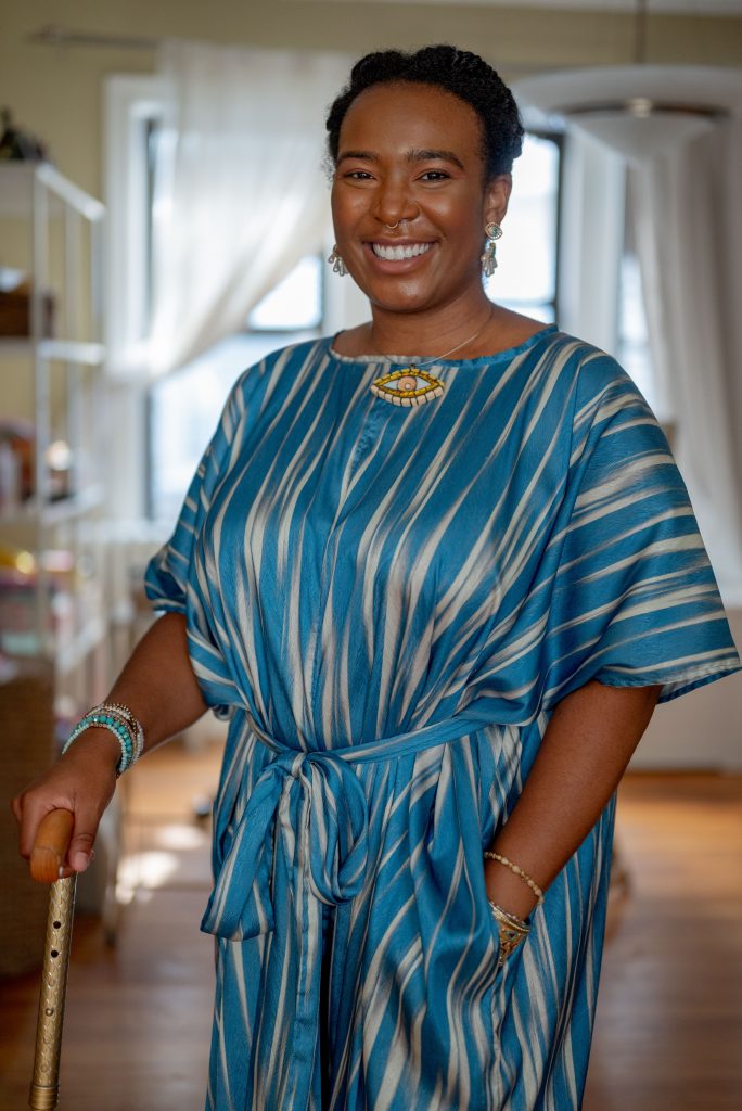 Image: Portrait of Perry standing up facing the camera with a smile. Perry is wearing a blue and white silk jumpsuit with a necklace in the shape of an eye. Photo by Tonal Simmons. 