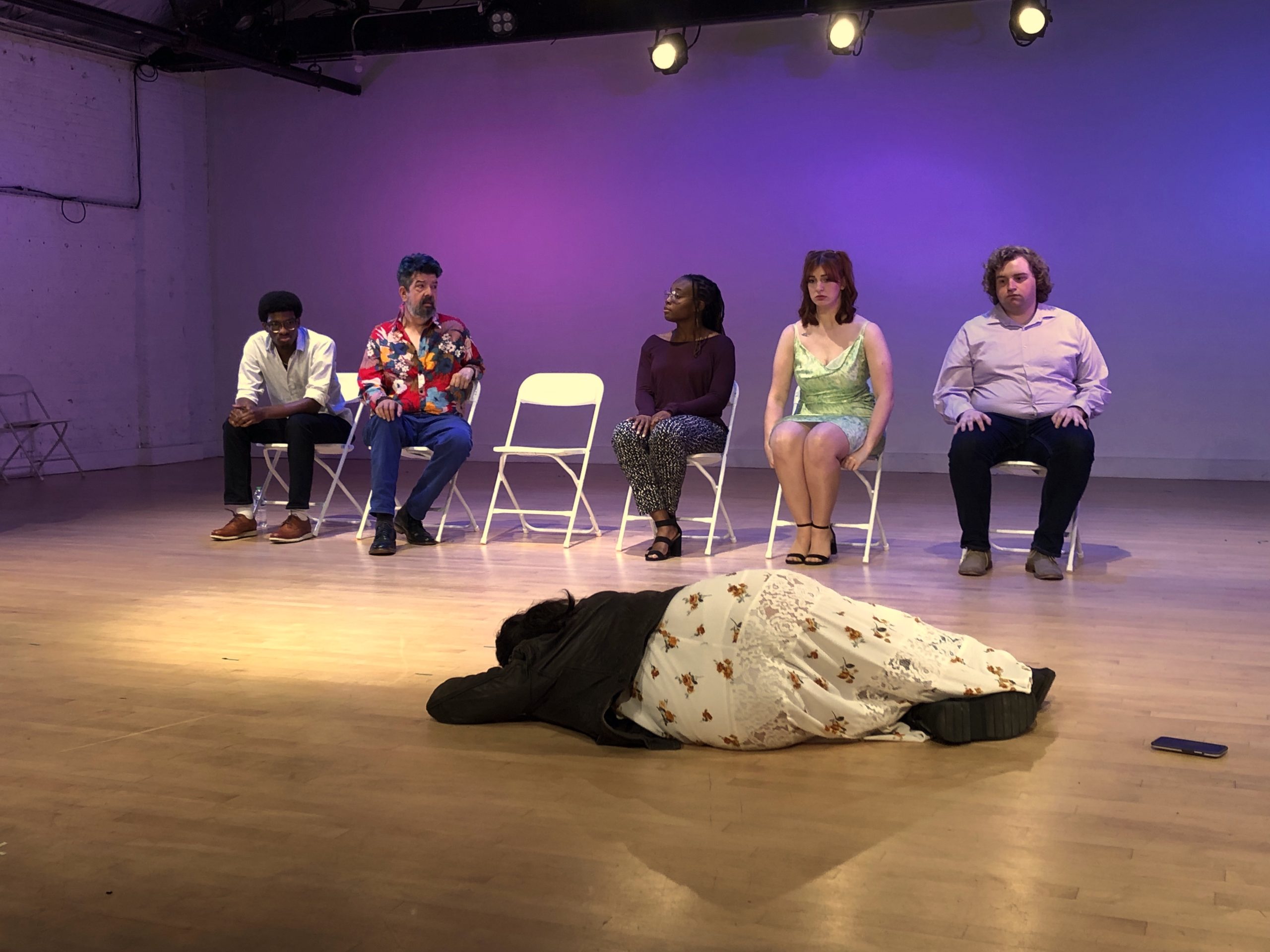 Image: Seated in a row on white folding chairs are Malik Middleton, Donaldson Cardenas, an empty chair, then OOshene Fox, Megan Kueter, and Michael Oakes. Lying on the light wooden floor before all of them is Haley Bolithon, facing towards the rest. The back is lit purple and pink. Photo by Hugh Graham.