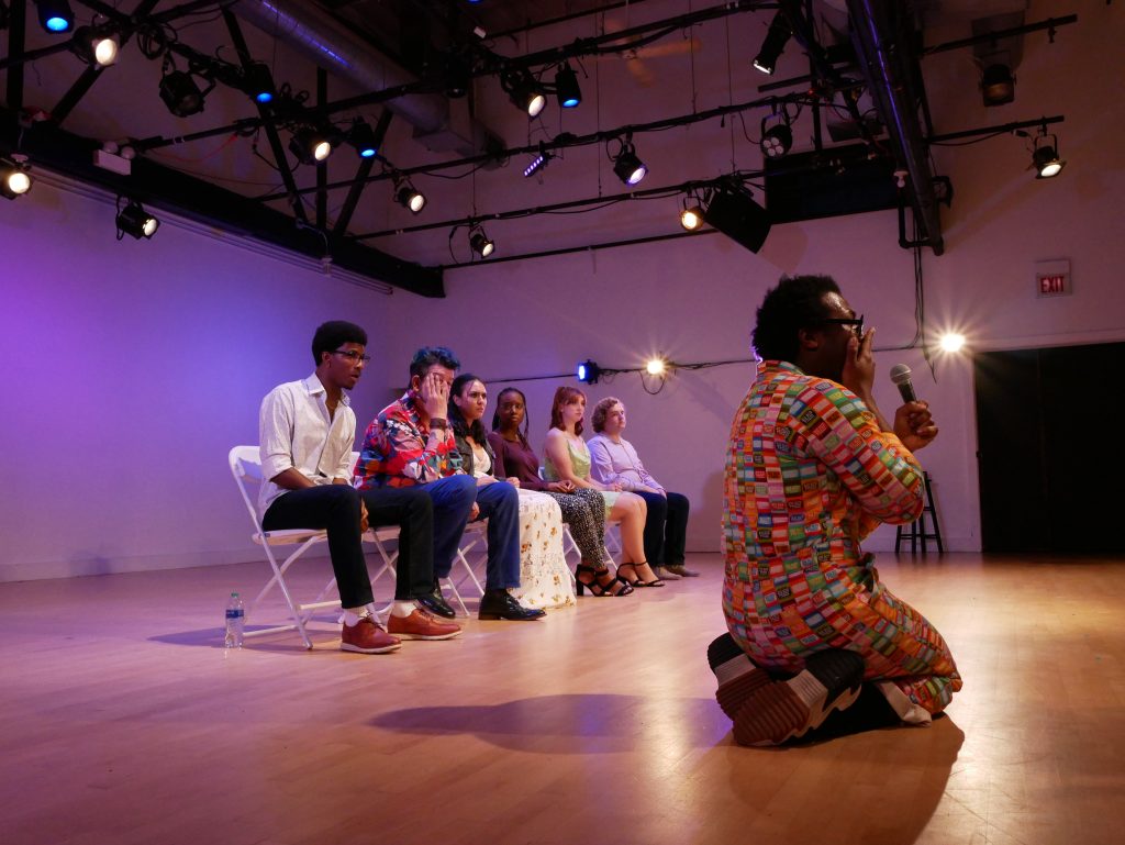 Image: Six people seated in white folding chairs. In foreground is a person on their knees holding a microphone and one hand to their mouth. Photo by Hugh Graham.