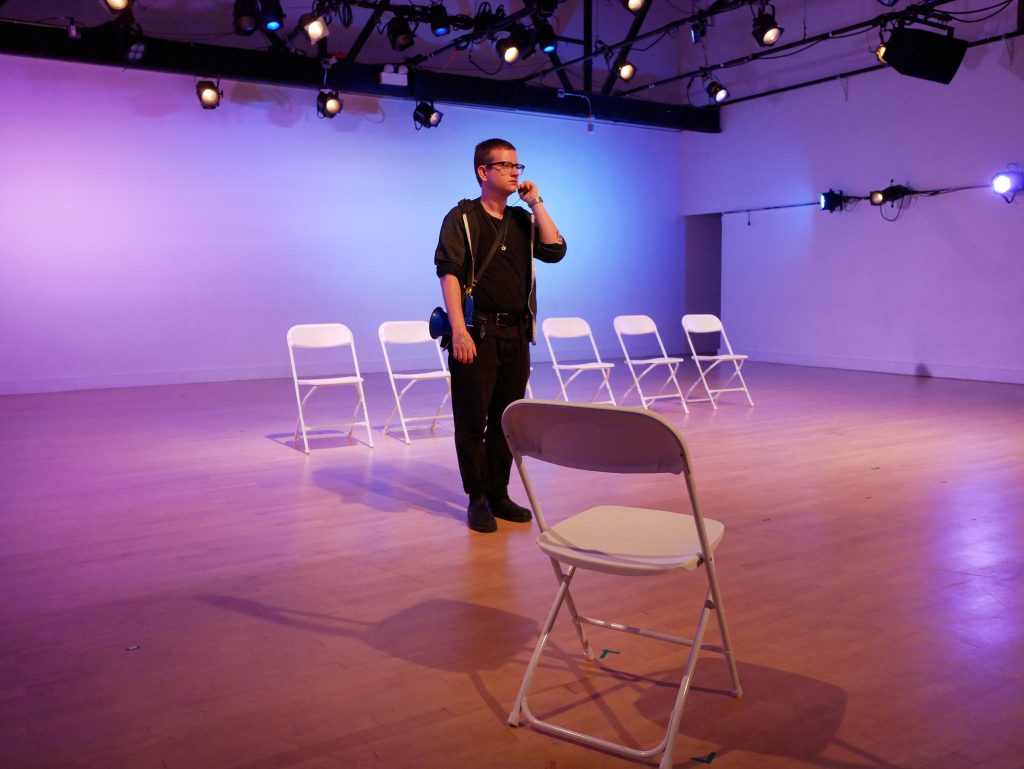 Image: A single figure standing centerstage with six white chairs behind them and one chair in front of them. A camera hangs around their chest. On the walls and ceiling are lights. Photo by Hugh Graham.
