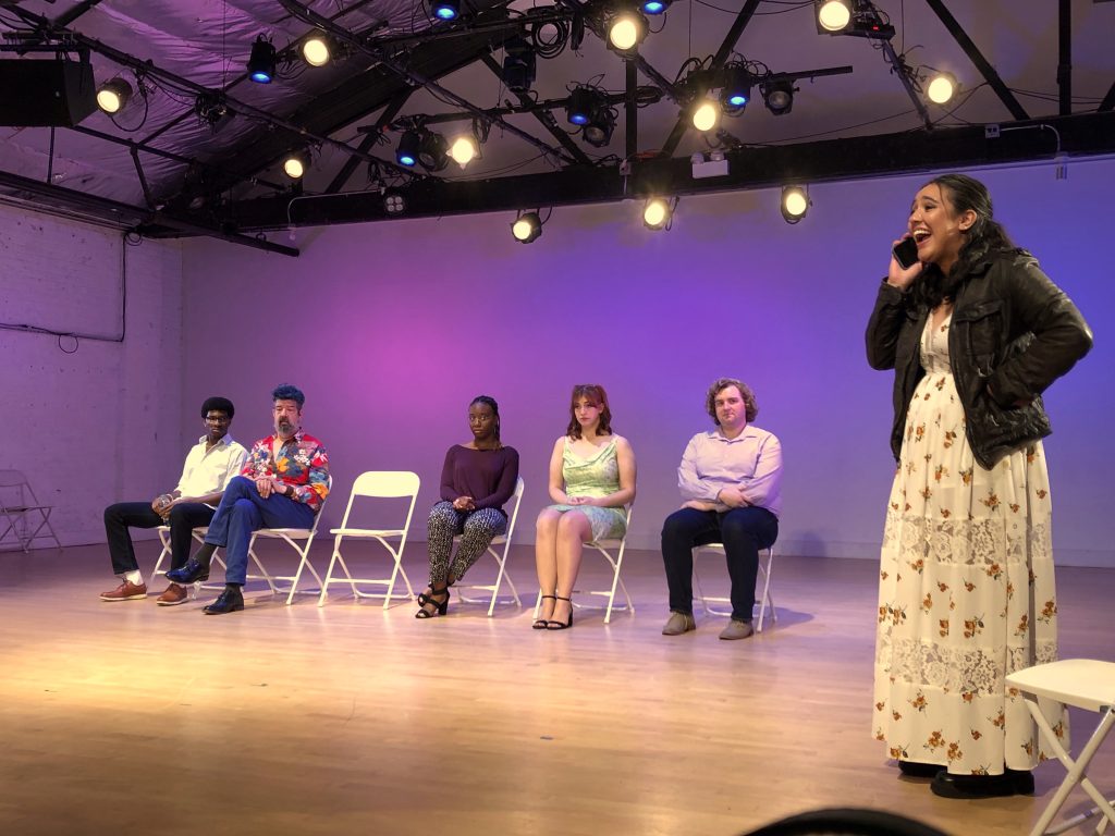 Image: Five people seated in a row on white folding chair. One chair is empty; the back wall is lit purple and pink. They all look at a person wearing a dress and a leather jacket smiling and holding a phone to their ear. Photo by Hugh Graham.