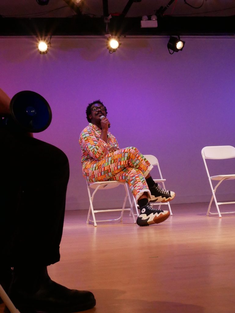 Image: Centered is a person seated, legs crossed, in a white folding chair, holding a mic. They lean back slightly, wearing converse shoes and a multi-colored gridded jumpsuit.. Beside them are two empty white chairs. On the left is a large silhouette of somebody's leg and camera lens. Photo by Hugh Graham.