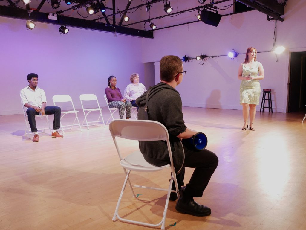 Image: Five people on stage. One is seated in the foreground facing a person in a green dress and heels to the right. On the left side of the stage are three people seated on white folding chairs and two empty chairs. Photo by Hugh Graham.