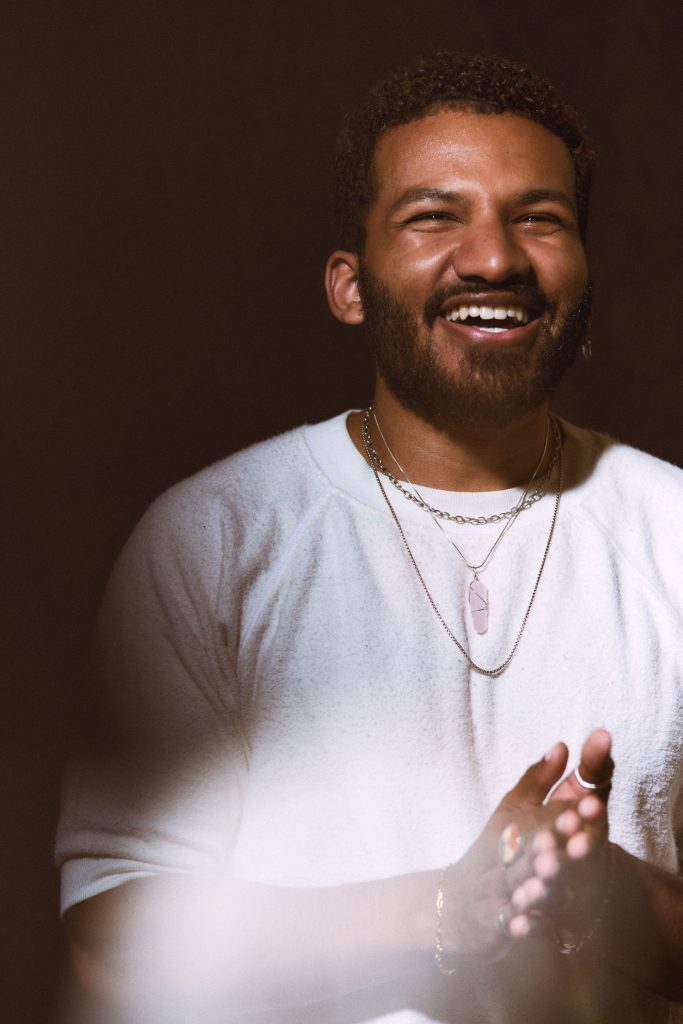Image: Rich Robbins is photographed against a black background. Robbins has short, curly hair and is wearing a white crewneck sweater, several thin necklaces, and a couple bracelets. He looks up smiling with his hands together. Photo by sarah joyce.