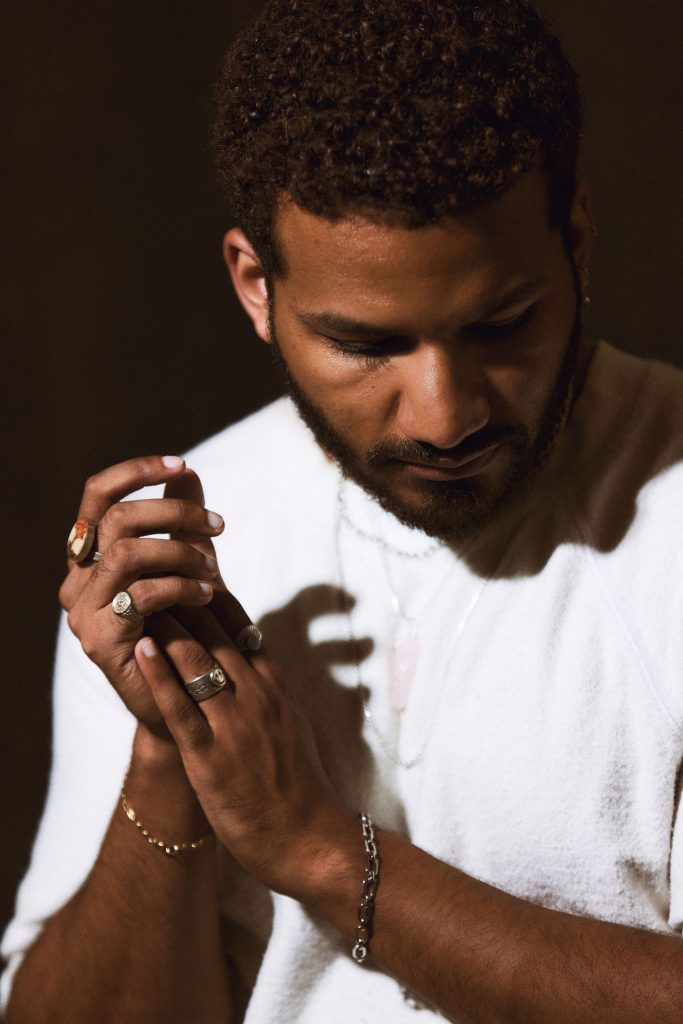 Image: Rich Robbins is photographed against a black background. Robbins has short, curly hair and is wearing a white crewneck sweater, several thin necklaces, and a couple bracelets. He looks down with his hands together. Photo by sarah joyce.