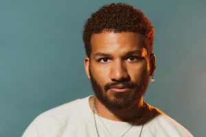 Rich Robbins is photographed against a muted gray background. Robbins is a Black man with short, curly hair. He is wearing a white crewneck sweater, several thin necklaces, and has a few earrings on his left ear.