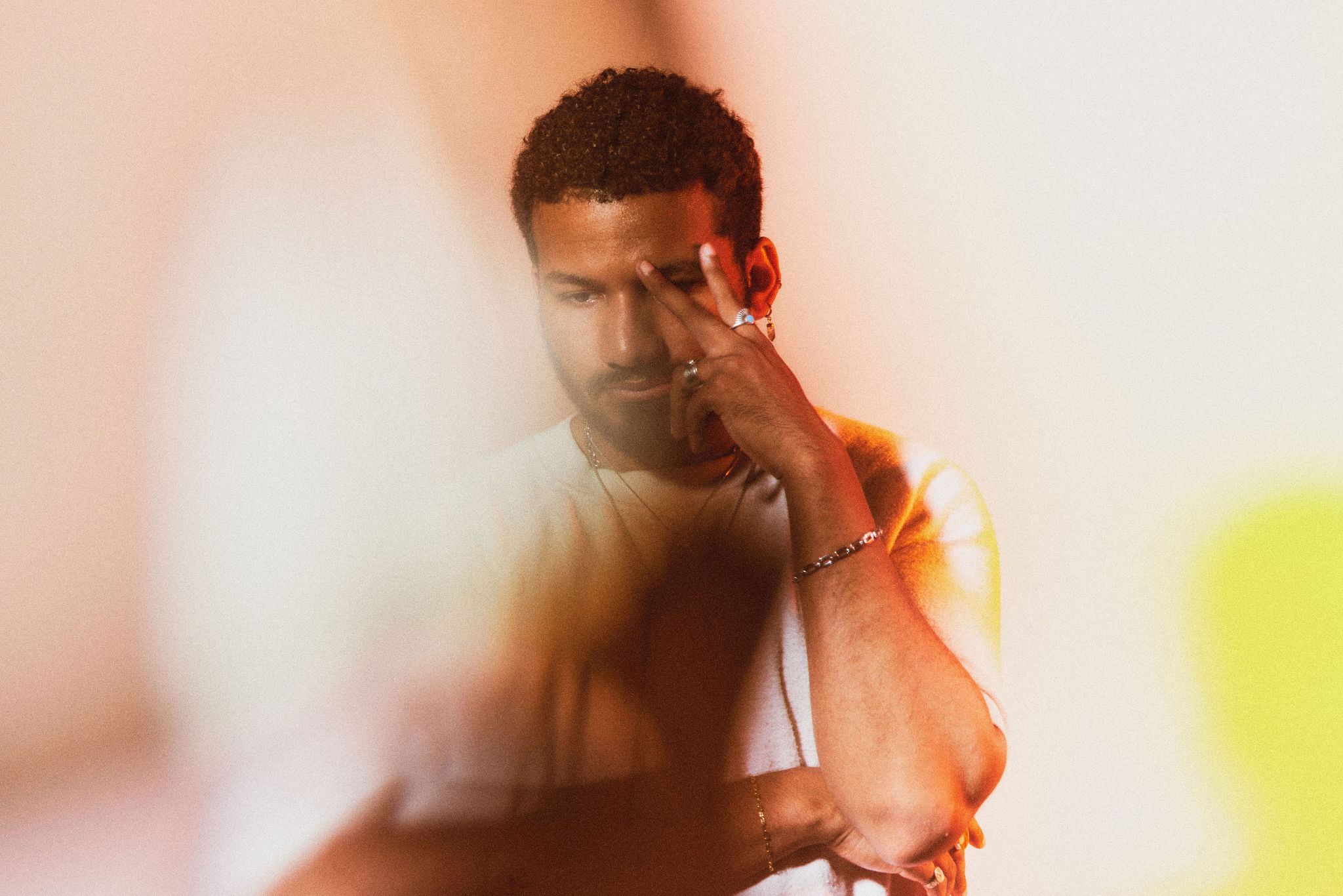 Image: Rich Robbins is photographed against a pale pink background. Robbins has short, curly hair and a beard. He is wearing a white crewneck sweater, and is casting his gaze downwards. His right arm is crossing his chest, supporting his left arm, which is raised, his hand making a V shape in front of his eye. Photo by sarah joyce.