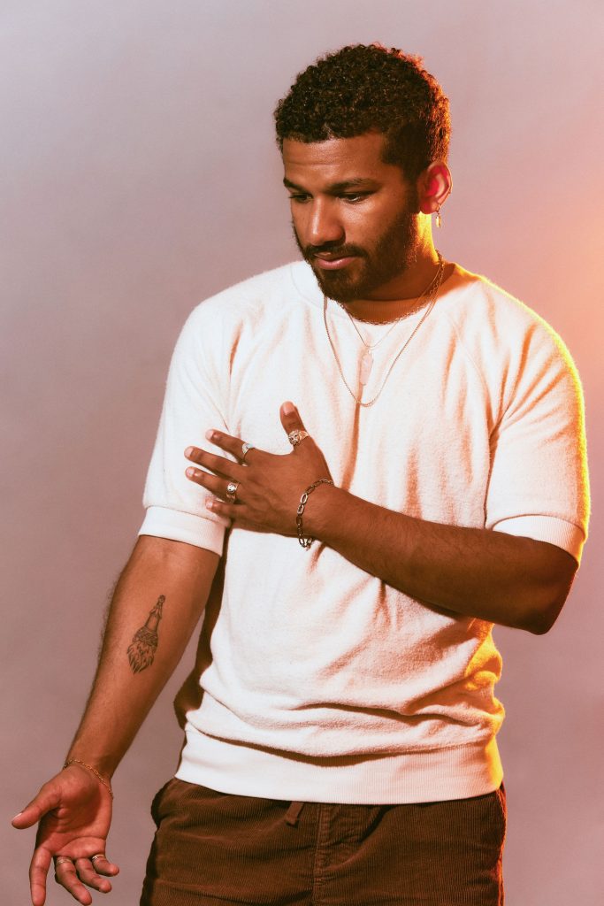 Image: Rich Robbins is photographed against a hazy pink and orange background. Robbins has short, curly hair and is wearing a white crewneck sweater, several thin necklaces, and has a few earrings on his left ear. He stands with one arm crossed over his chest and the other extended down. Photo by sarah joyce.