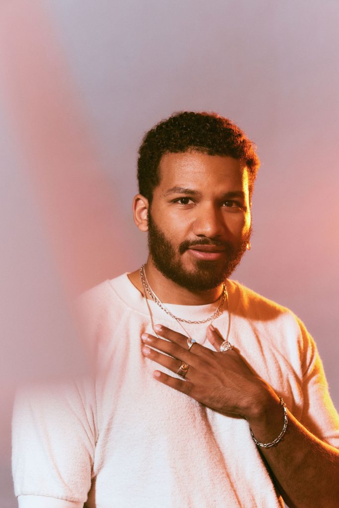 Image: Rich Robbins is photographed against a hazy pink and orange background. Robbins has short, curly hair and is wearing a white crewneck sweater, several thin necklaces, and has a few earrings on his left ear. He has one hand on his chest. Photo by sarah joyce.