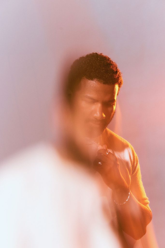 Image: Rich Robbins is photographed against a hazy pink and orange background. Robbins has short, curly hair and is wearing a white crewneck sweater, several thin necklaces, and has a few earrings on his left ear. He has one arm bent upward towards his face. The photo has an ethereal, hazy effect. Photo by sarah joyce.