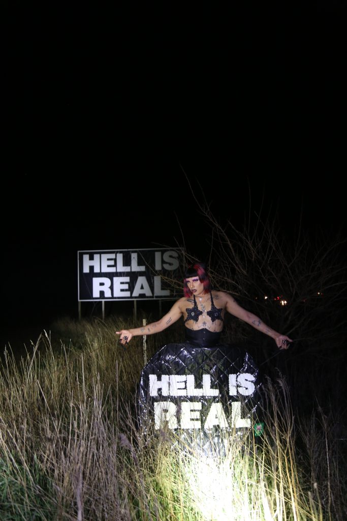 Image: Celeste stands in front of a billboard that reads “Hell is Real” along a highway in Indiana. She is wearing a black, star-shaped silicone bra, a quilted skirt adorned with the same statement, and a black and magenta bob wig. Photo by Ruby Que.