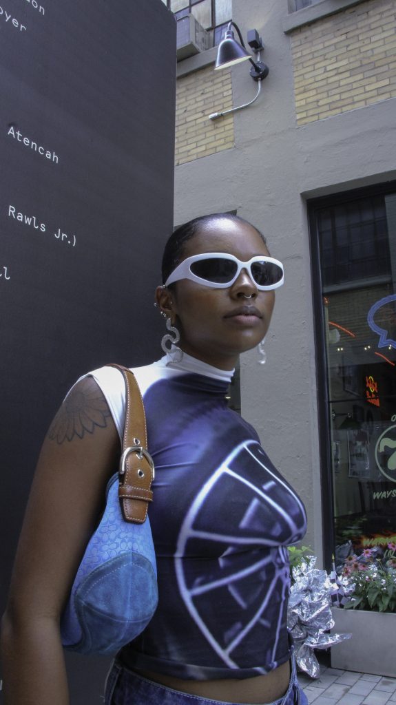 Image: Arria @lilbabyog. Aria pairs a fitted spandex top with a large tire graphic on it with white sunglasses, white squiggly earrings, white, black, and purple bracelets, denim capris, and a blue Coach shoulder bag. Photo credit: Salyse.