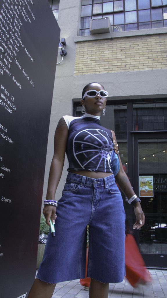 Image: Arria @lilbabyog. Aria pairs a fitted spandex top with a large tire graphic on it with white sunglasses, white squiggly earrings, white, black, and purple bracelets, denim capris, and a blue Coach shoulder bag. Photo credit: Salyse.