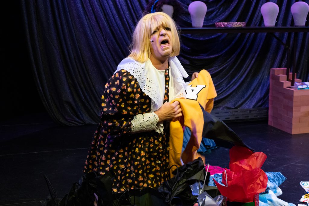 Image: Benjamin Larose on stage in a choppy blond wig and dated floral dress with a large white lace collar and cuffs. Kneeling on the floor and grasping a yellow sweatshirt, the character is wailing exaggeratedly. Photo courtesy of the artist, Benjamin Larose.