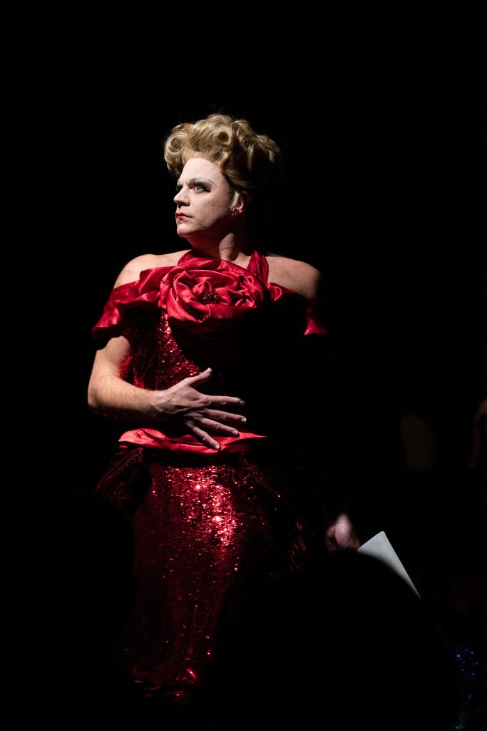 Image: Benjamin Larose, in the spotlight on stage in full drag wearing a sparkly red dress and blonde wig fashioned in a curly updo. Photo courtesy of the artist, Benjamin Larose.