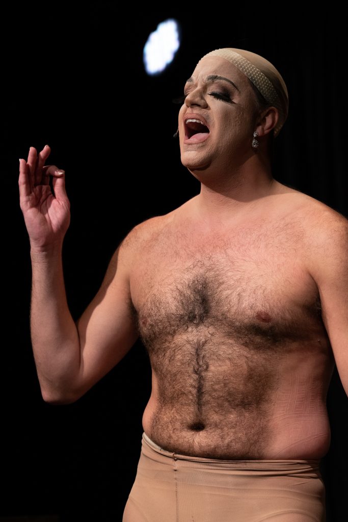 Image: Artist Benjamin Larose on stage in full makeup with diamond earrings and nude hair cap. Shirtless, the artist is singing passionately with his eyes closed. Photo courtesy of the artist, Benjamin Larose.