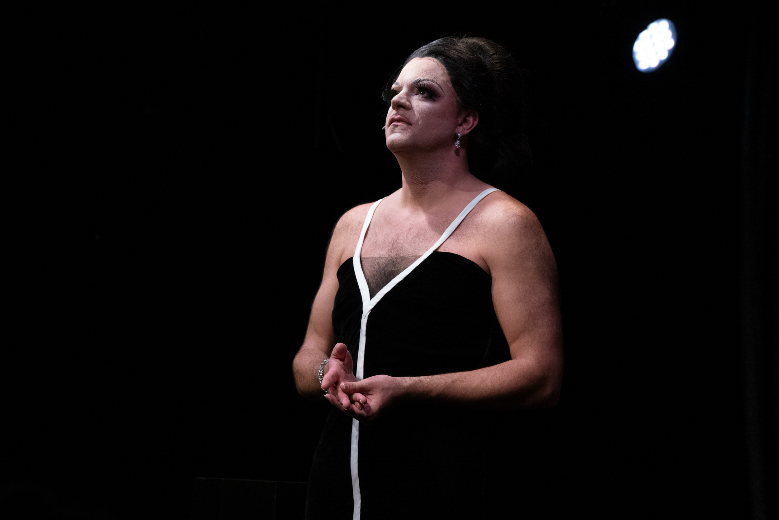 Image: Benjamin Larose onstage. Grasping his hands pensively, he looks upwards towards the spotlight shining down on him. Photo courtesy of the artist, Benjamin Larose.