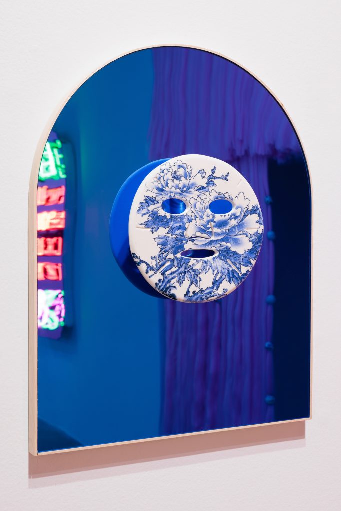 Images: Jennifer Ling Datchuk, Flawless (Blue), 2022. Porcelain, decals from Jingdezhen, China, mirror, 16 x 20 x 3". A blue-tinted mirror with a mask-like face in the center. The mask is white with blue floral shapes. Image courtesy of the artist and Bemis Center for Contemporary Arts. Photography by Colin Conces.