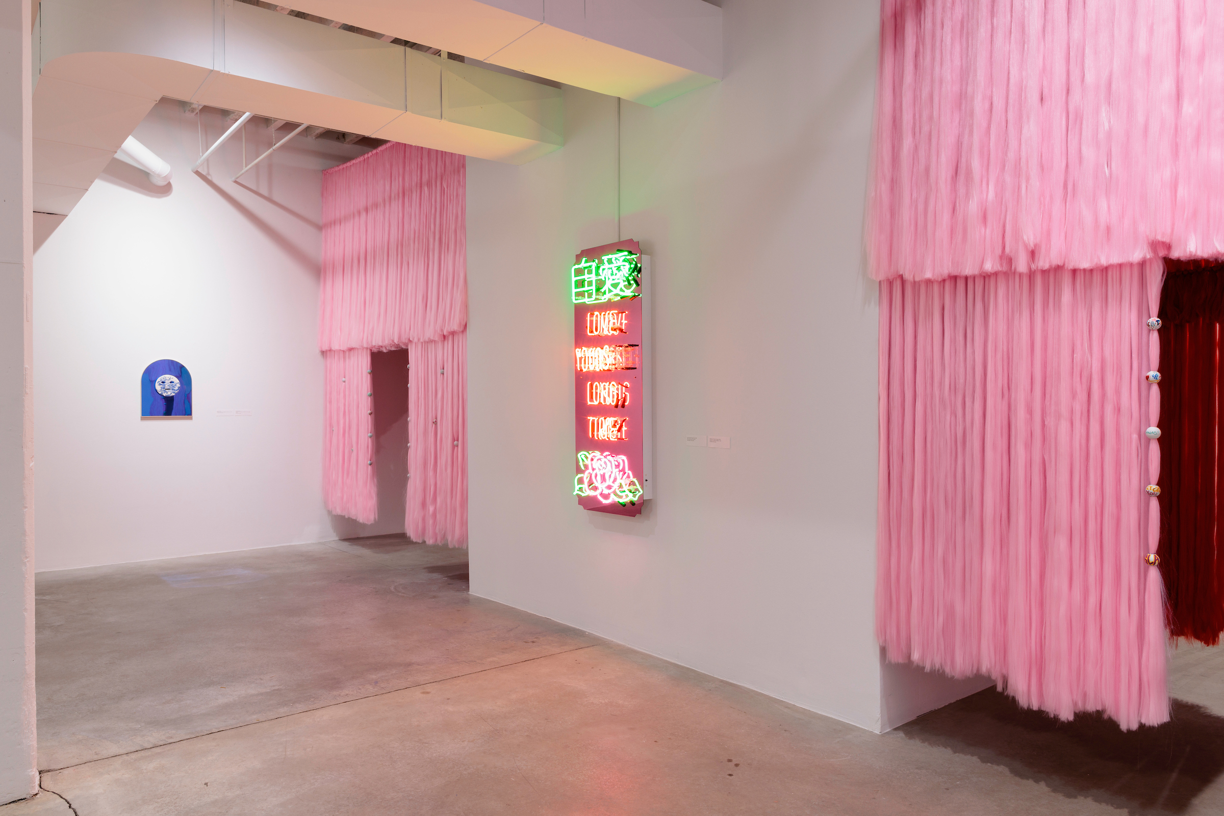 Image: Installation view of Jennifer Ling Datchuk: Eat Bitterness, 2023. In the center we can see Jennifer Ling Datchuk's piece Love Yourself Long Time, a pink-tinted mirror with neon letters that say "LOVE YOURSELF LONG TIME" in English and Chines, with a rose at the bottom. There are pink curtains on either side of the piece. Courtesy of Bemis Center for Contemporary Arts. Photography by Colin Conces.