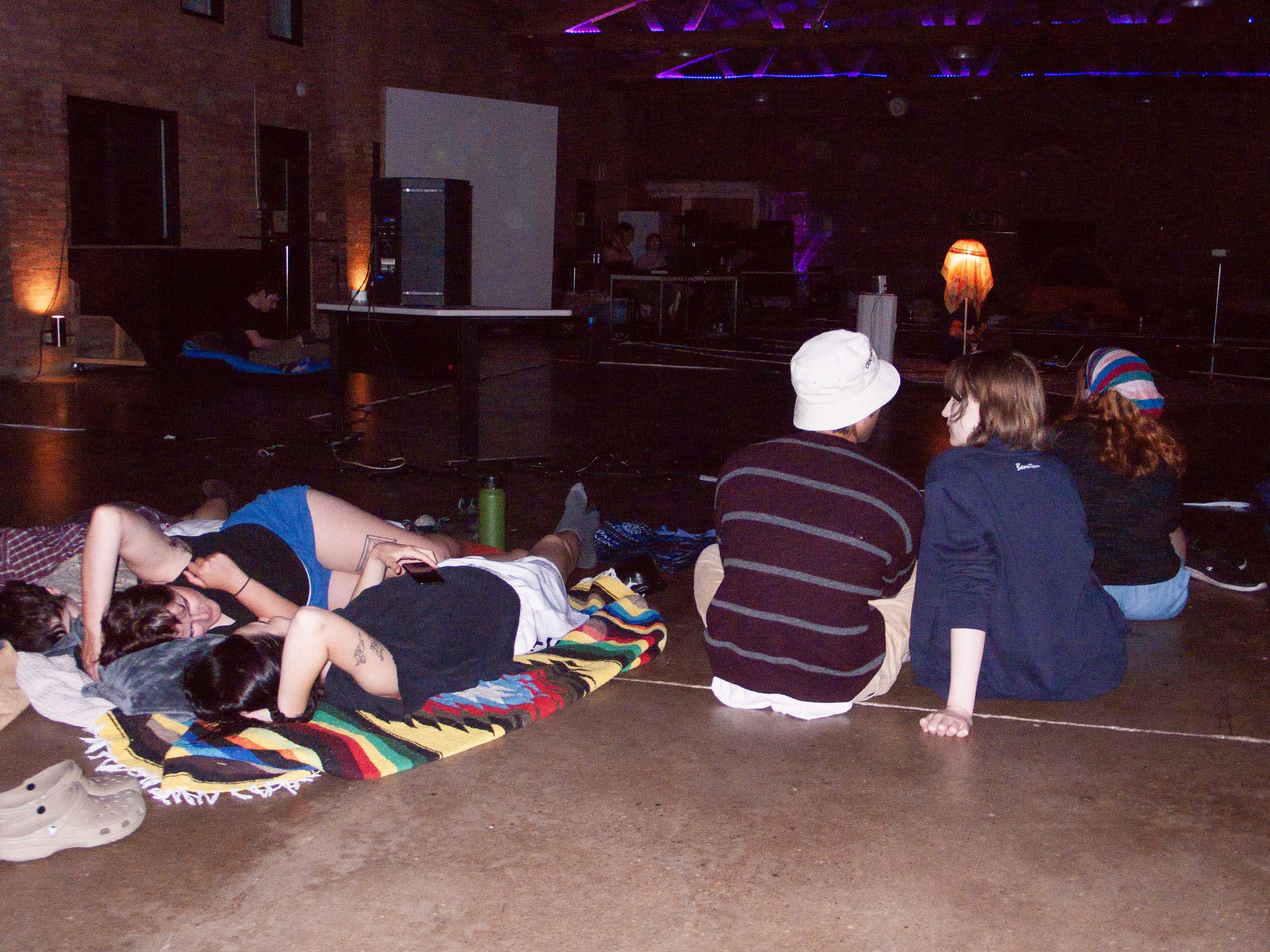 Image: Image: The crowd sitting and laying in a dark room on a polished concrete floor at StretchMetal's Drone Sleepover. They face away from the camera, and towards a stage with a large speaker, other sound equipment, and a floor lamp with a large, translucent sheet over it. Image courtesy of StretchMetal.