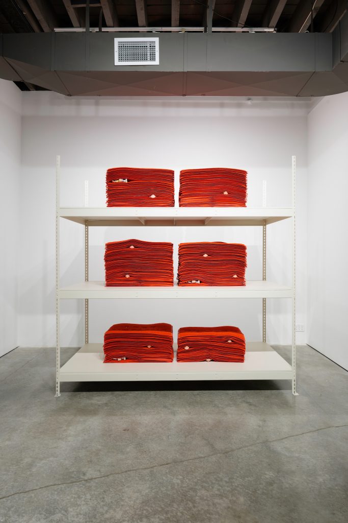 Left Image: Jennifer Ling Datchuk. Live to Die, 2019. Custom red welcome mats and porcelain, varied dimensions. Stacks of red welcome mats can be seen on a white shelving unit. In between some of the mats are small, porcelain figurines. Originally commissioned by the Black Cube Nomadic Museum. Image courtesy of the artist and Bemis Center for Contemporary Arts. Photography by Colin Conces. 