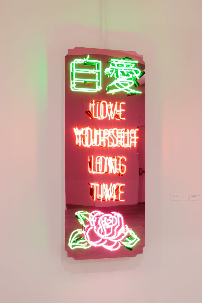 Image: Jennifer Ling Datchuk. Love Yourself Long Time, 2021. Mirrored acrylic and neon, 60 x 24 x 5". A pink-tinted mirror with neon letters that say "LOVE YOURSELF LONG TIME" in English and Chines, with a rose at the bottom. Image courtesy of the artist and Bemis Center for Contemporary Arts. Photography by Colin Conces.