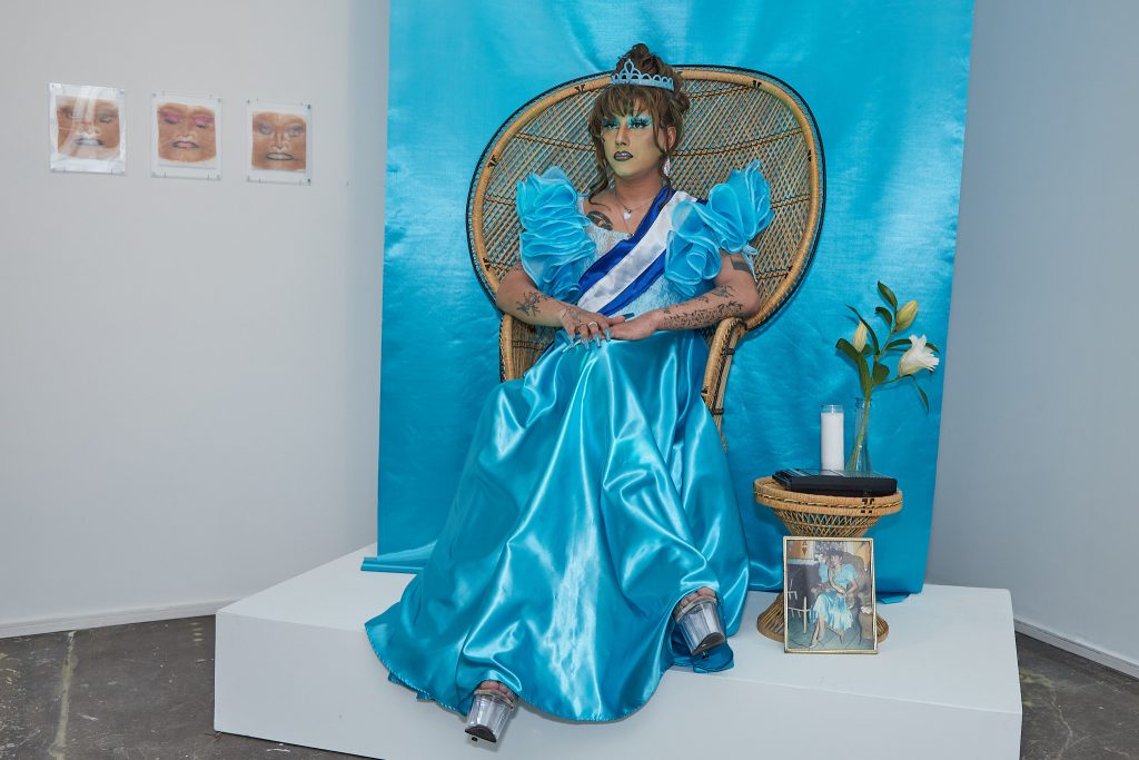 Image: Ále Campos performs as Celeste at the Newcity Breakout Artists 2023 Exhibition. Celeste sits on a throne-like rattan arm chair in front of a blue satin background. She is wearing pleaser heels, a blue satin dress with ruffled shoulders, a white and blue sash, and a ceramic tiara. A photo of her mother wearing a similar dress is presented on the plinth beside her, as well as a tape player, a candle, and a single flower stem. Three of their framed makeup wipes are hung on the wall to their right. Photo by Ashley Baranczyk.