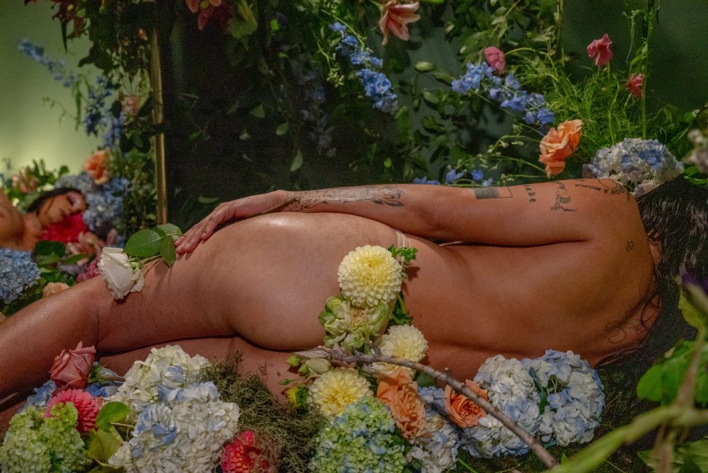 Image: A still shot of “At Rest,” a collaboration between Vince Phan and Ále Campos, presented by Jude Gallery. A figure sleeps in a bed of flowers and moss. They are turned away from the camera, but their face is visible in the reflection of a mirror placed behind them. Photo by Tonal Simmons.
