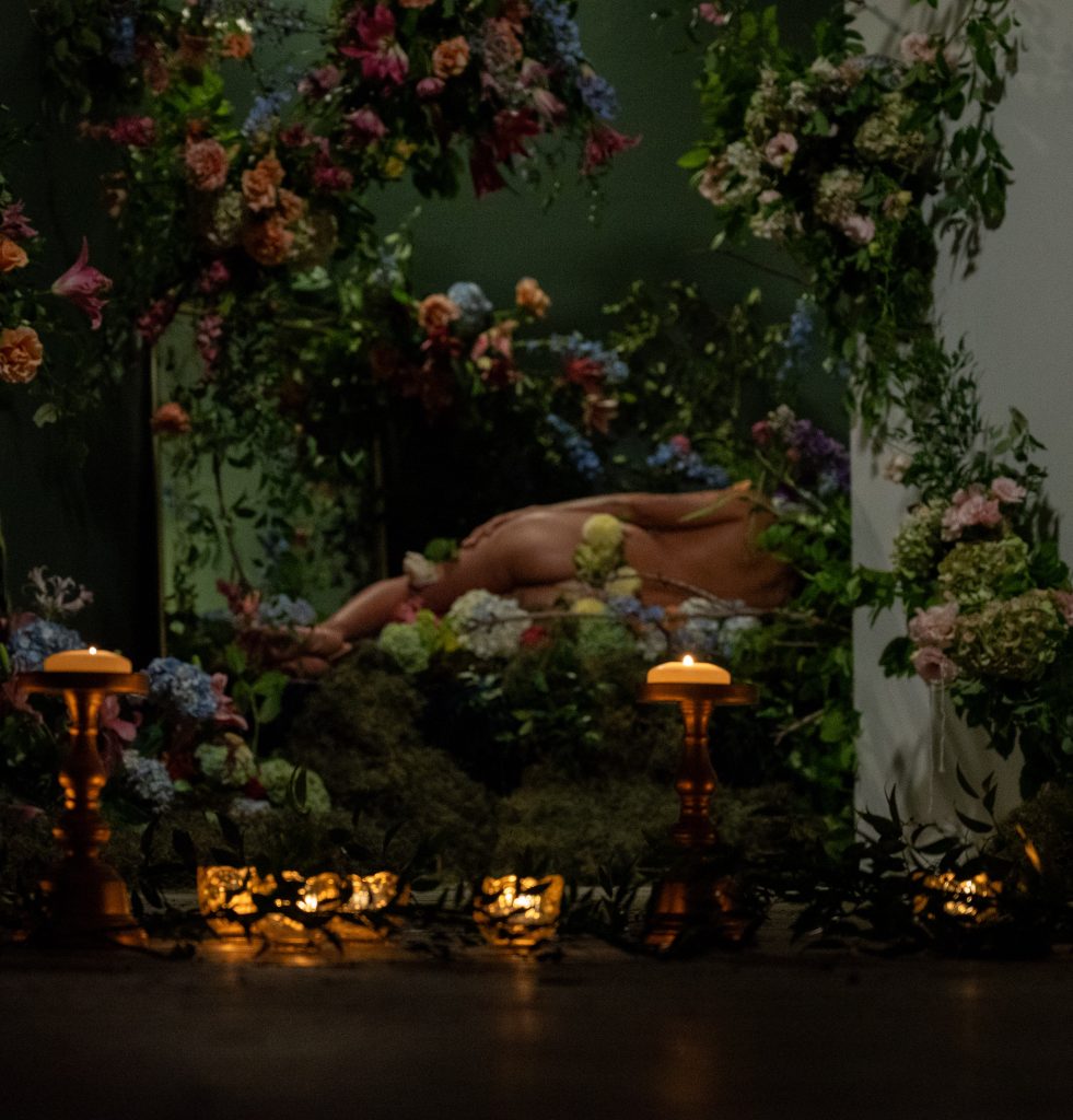 Image: A still shot of “At Rest,” a collaboration between Vince Phan and Ále Campos, presented by Jude Gallery. A naked figure reclines on a bed of moss and flowers. They are turned away from the camera. Small tealight candles dot the foreground along with small, leafy twigs. Photo by Tonal Simmons.