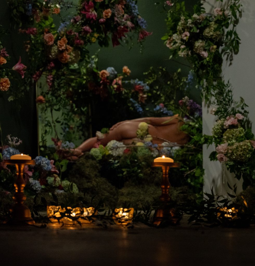 Imagen: Una fotografía de “At Rest / En Reposo”, una colaboración entre Vince Phan y Ále Campos, presentada por Jude Gallery. Una figura desnuda reclinada sobre una cama de musgos y flores. Está dando la espalda a la cámara. Pequeñas velas decoran el plano junto con pequeñas ramas de hojas. Crédito de imagen: Tonal Simmons