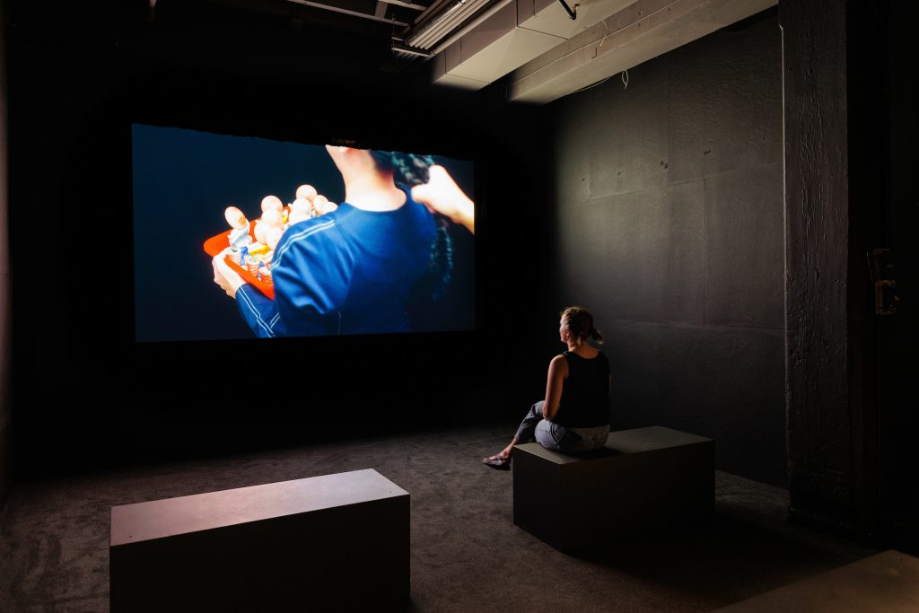 Image: Jennifer Ling Datchuk. Tame, 2021. Video and Synthetic Hair. A person sits on a bench in a black room watching the artist's video piece Tame. The still of the film shows a person pulling the artist's hair while she carries a basket filled with figurines. Image courtesy of the artist and Bemis Center for Contemporary Arts. Photography by Colin Conces.