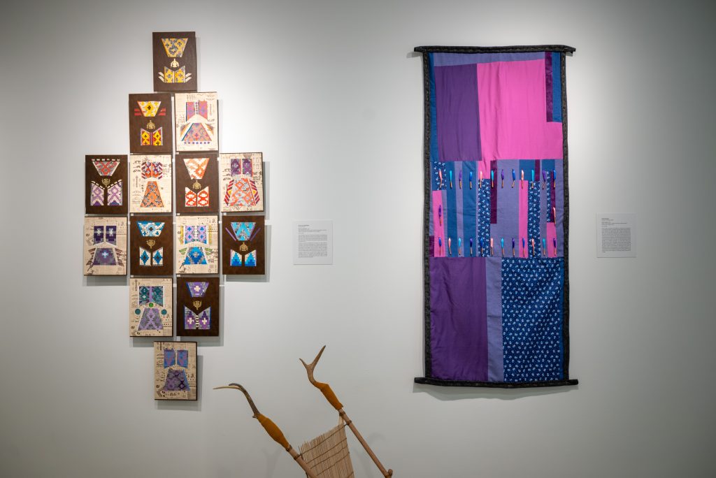 Image: Installation view of Native Futures. Artist Hattie Lee Mendoza's mixed media print work Cherokee Britannica (left) consists of 13 pieces arranged in a diamond shape. Her mixed media work uses colored pencils, paint swatches, and book pages in German, English, and Spanish. Jason Wesaw's textile piece titled Nanaw’igwan (In the Middle of It) (right) is made of fabric in shades of purple, pink, and blue. At the bottom center is the top half of the handmade stroller with antlers as handles by Noelle Garcia. Photograph by Tonal Simmons. 